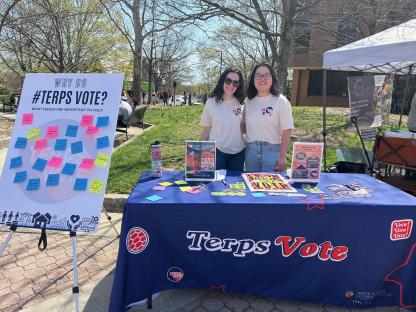 two students smiling and facing camera behind a table with a tablecloth reading "terpsvote"